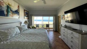 A hotel room with a bed, a television, a ceiling fan, a balcony with a view of the beach, and a painting on the wall. - Luxurious oceanfront apartment LMB2 (San José del Cabo)