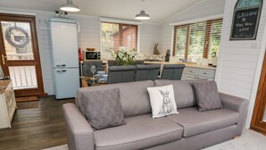 A modern living room with a grey sofa, a kitchen in the background, and a welcome sign on the wall. - Tree View Lodge (Windermere)