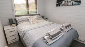 A bedroom with a bed, a window with blinds, a nightstand with a lamp, a small plant, and a painting on the wall. - Tree View Lodge (Windermere)