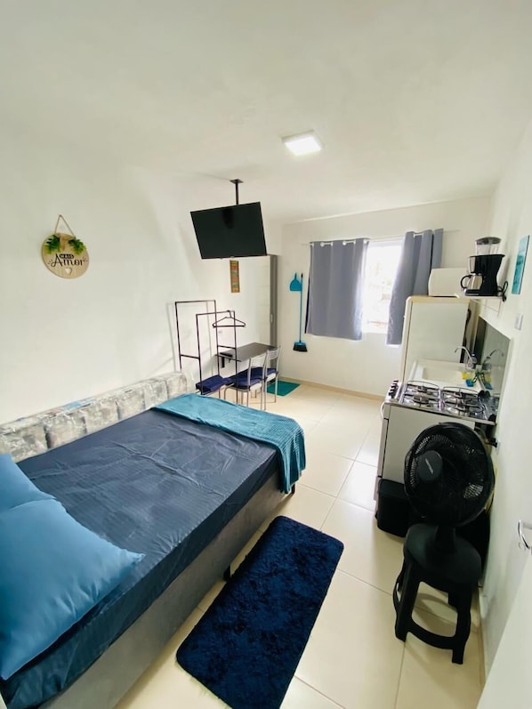 Een kleine, goed verlichte kamer met een bed, een bureau met een computer, een stoel, een klein tafeltje en een raam met gordijnen.