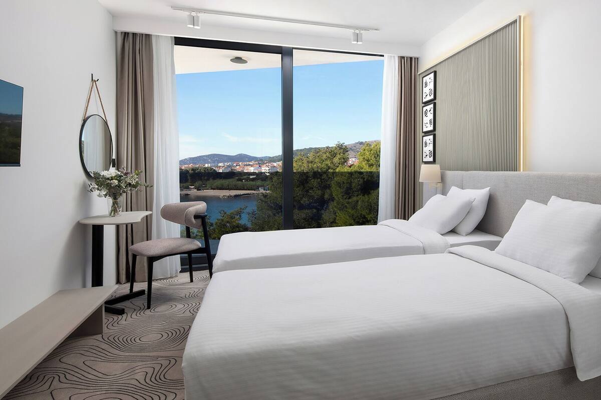 A hotel room with two beds, a chair, a table, and a view of a lake and cityscape.