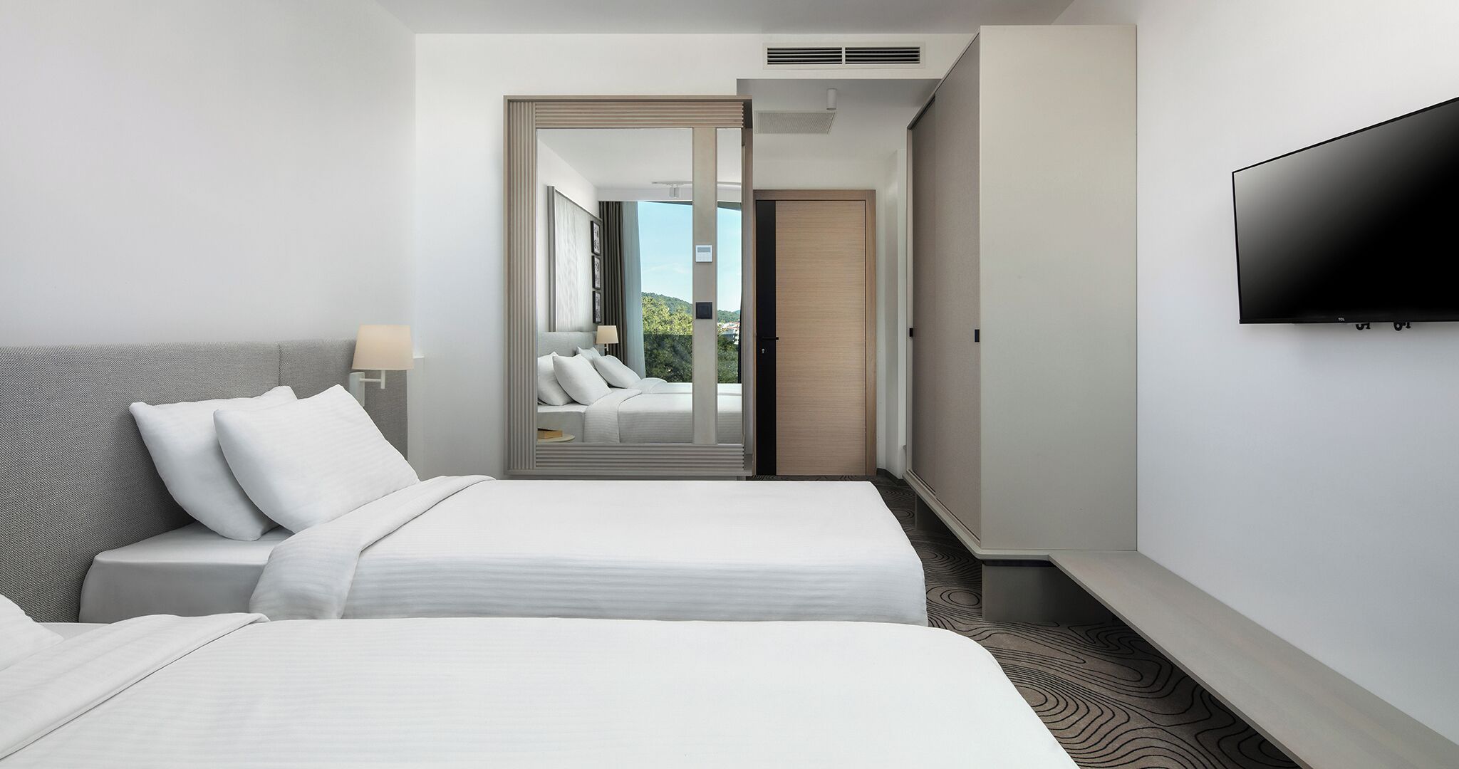 A hotel room with a bed, a TV, a mirror, and a view of the outdoors.