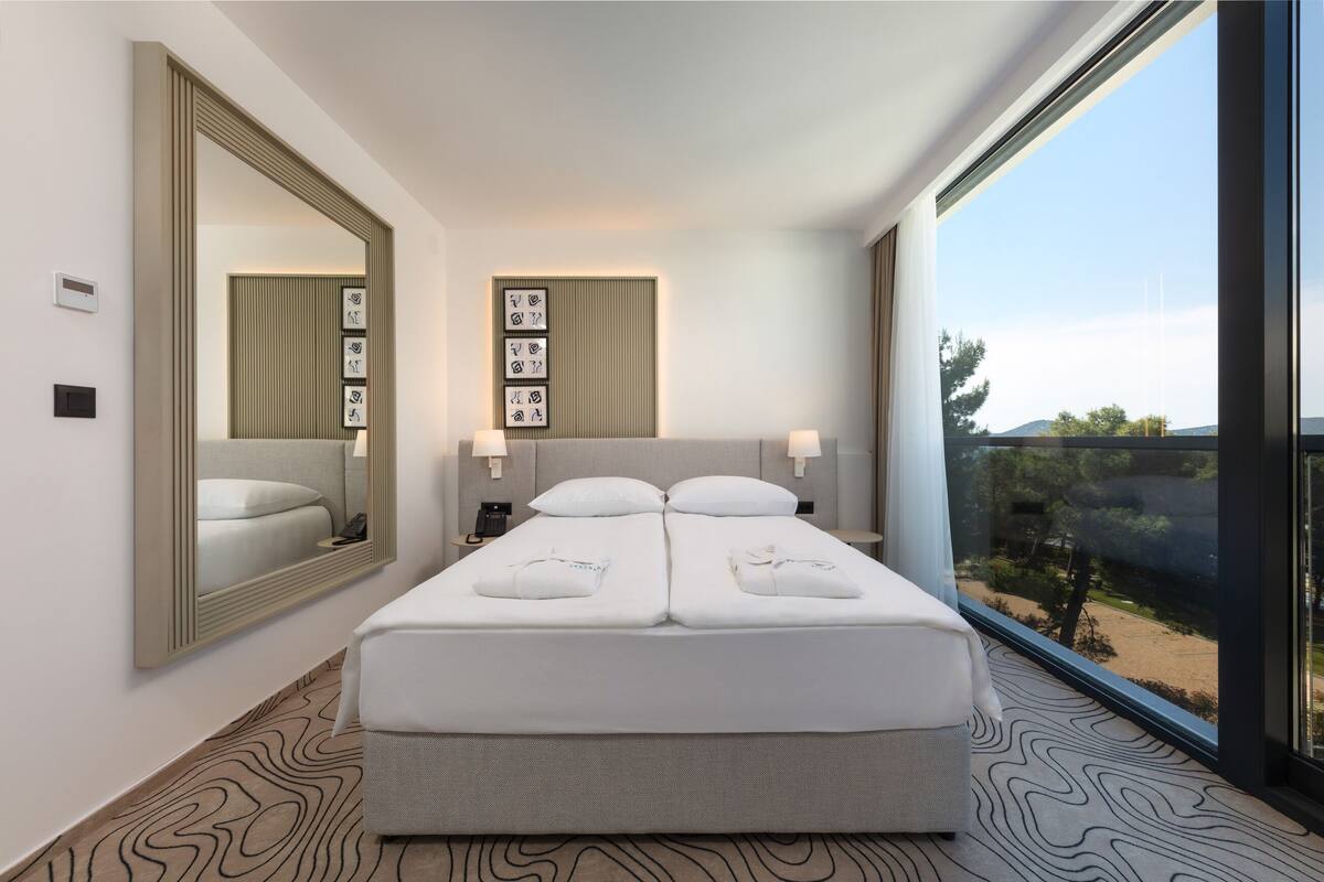 A modern hotel room with a large bed, a mirror, wall-mounted artwork, and a view of the outdoors.