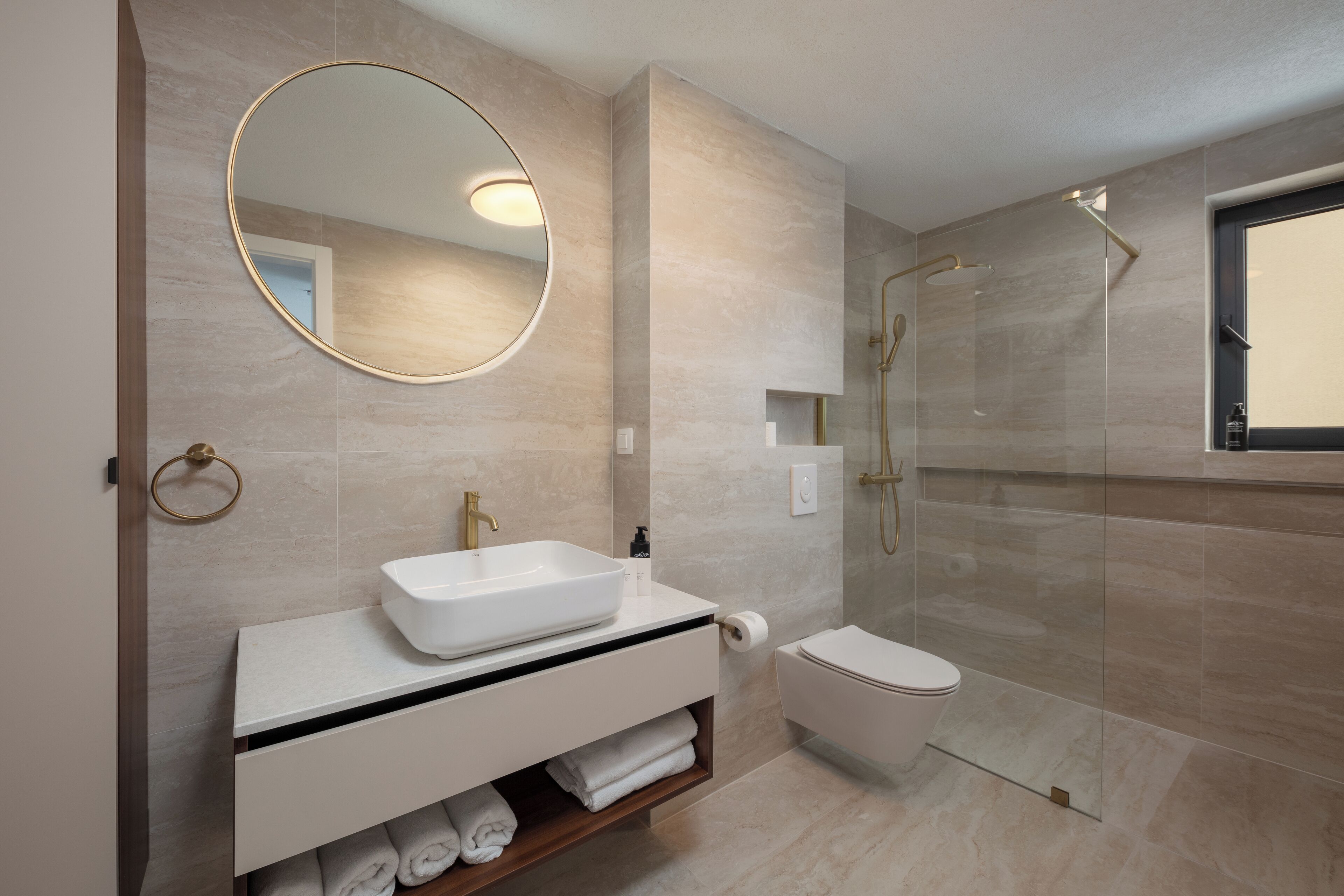 A modern bathroom with a large oval mirror, a white sink, a toilet, and a glass-enclosed shower area.