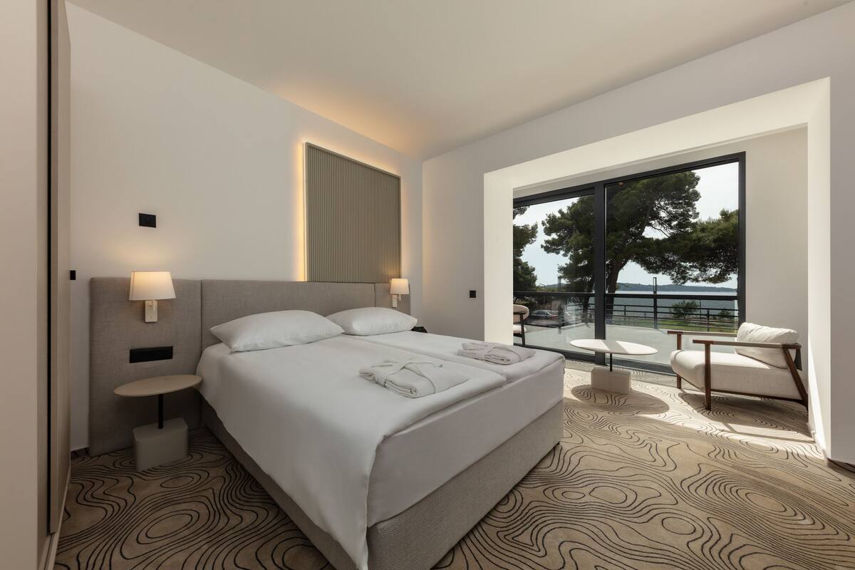 A modern hotel room with a large bed, a small round table, a chair, a balcony with a view of the sea, and a large window.