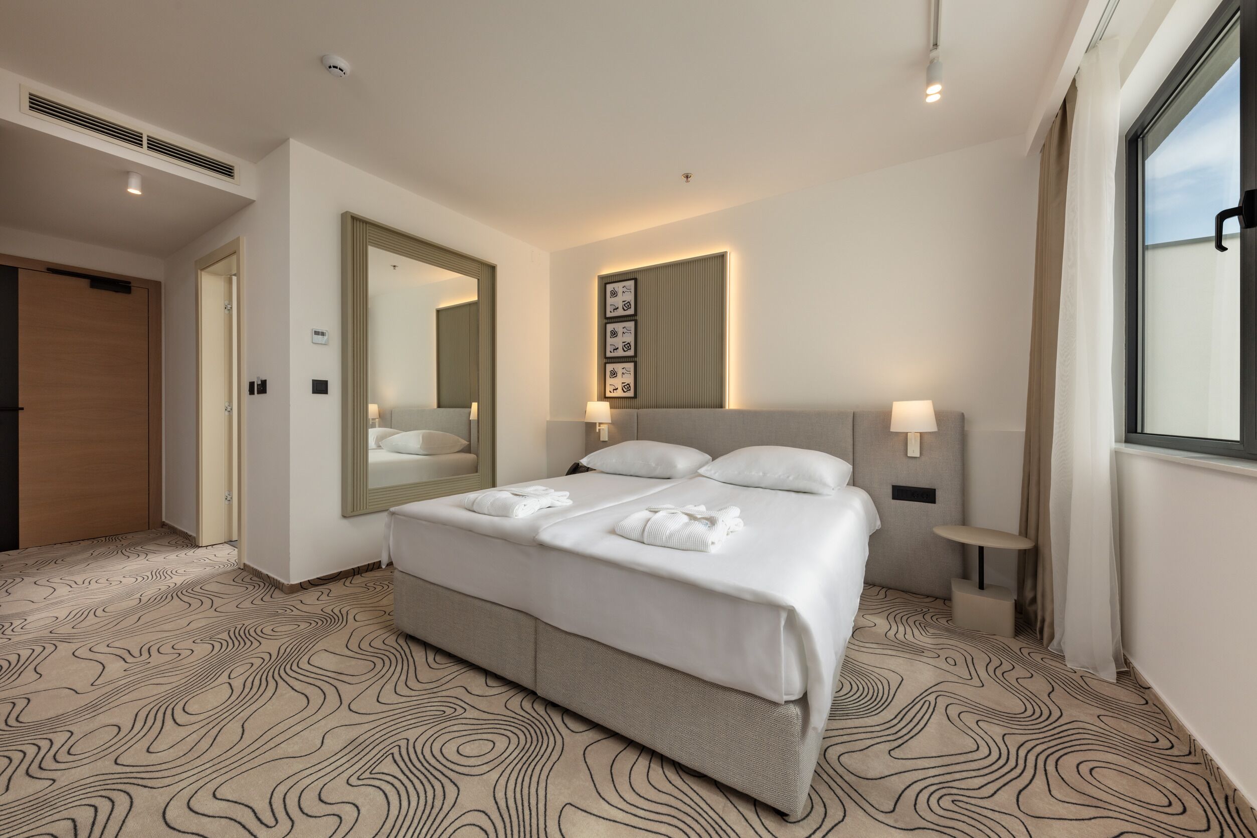 A hotel room with a large bed, a mirror, a bedside table with a lamp, and a window with curtains.