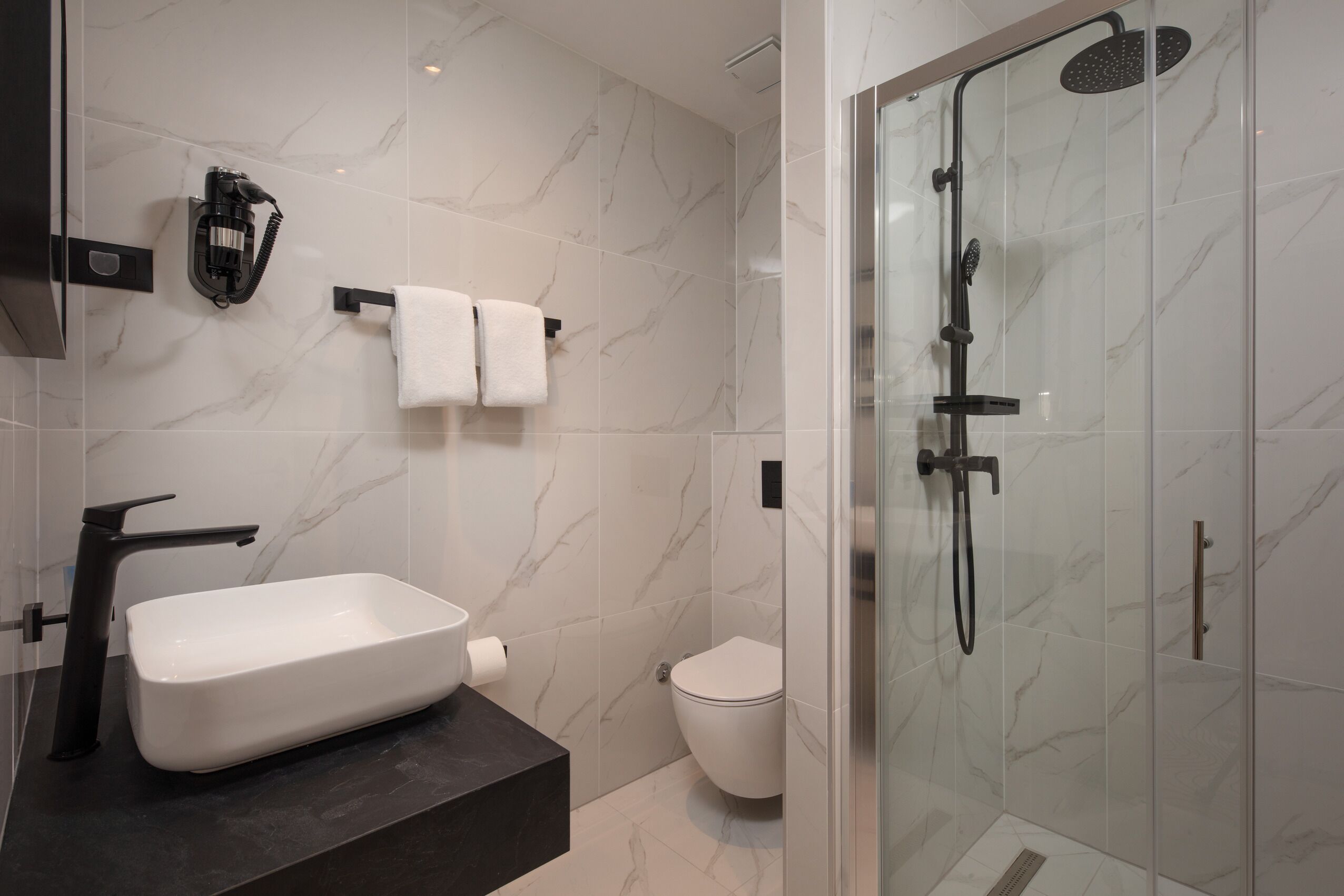 A modern bathroom with a marble backsplash, a white sink, a toilet, and a glass-enclosed shower area.