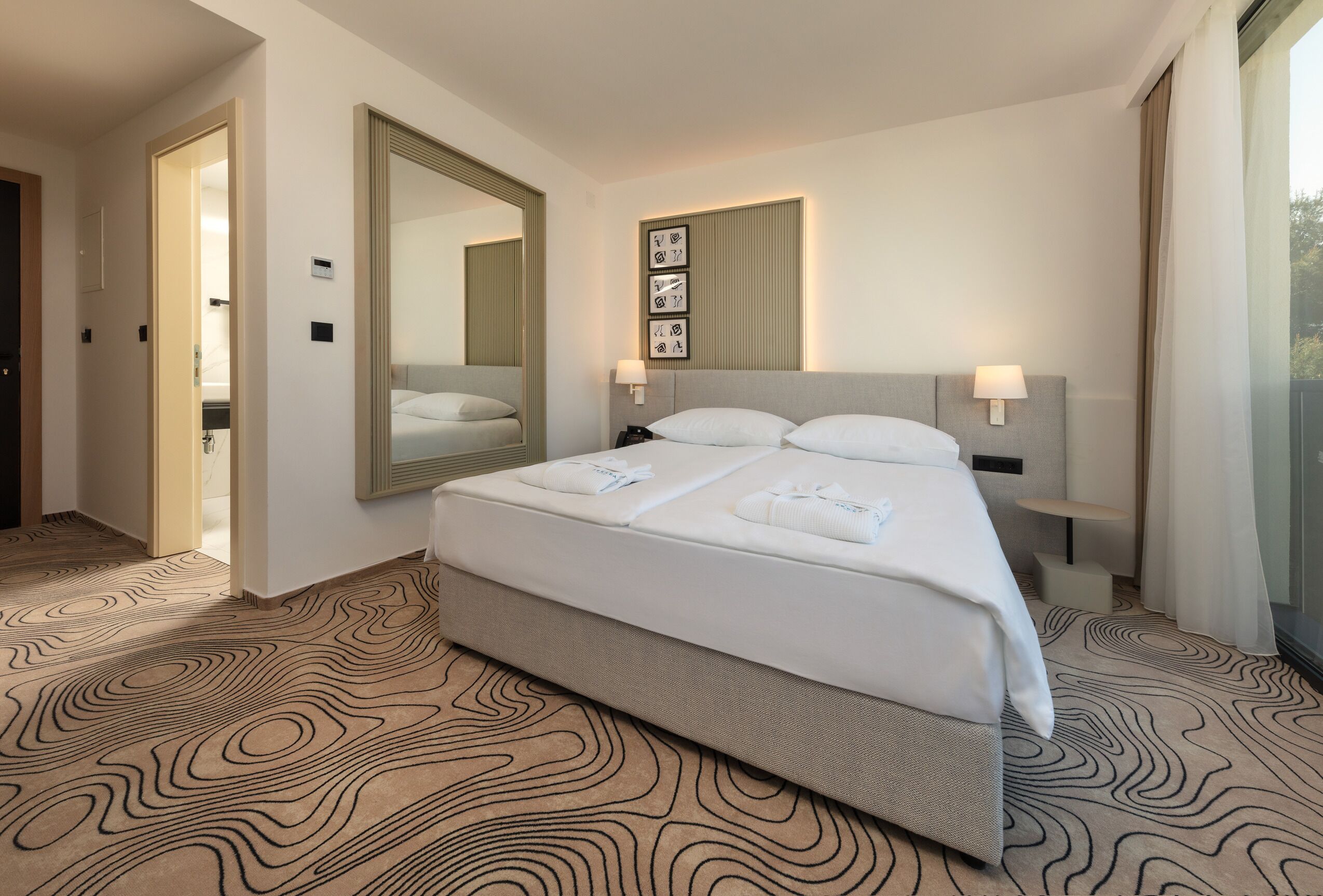 Quarto de hotel moderno com uma cama grande, um espelho, abajures e vista para o exterior.