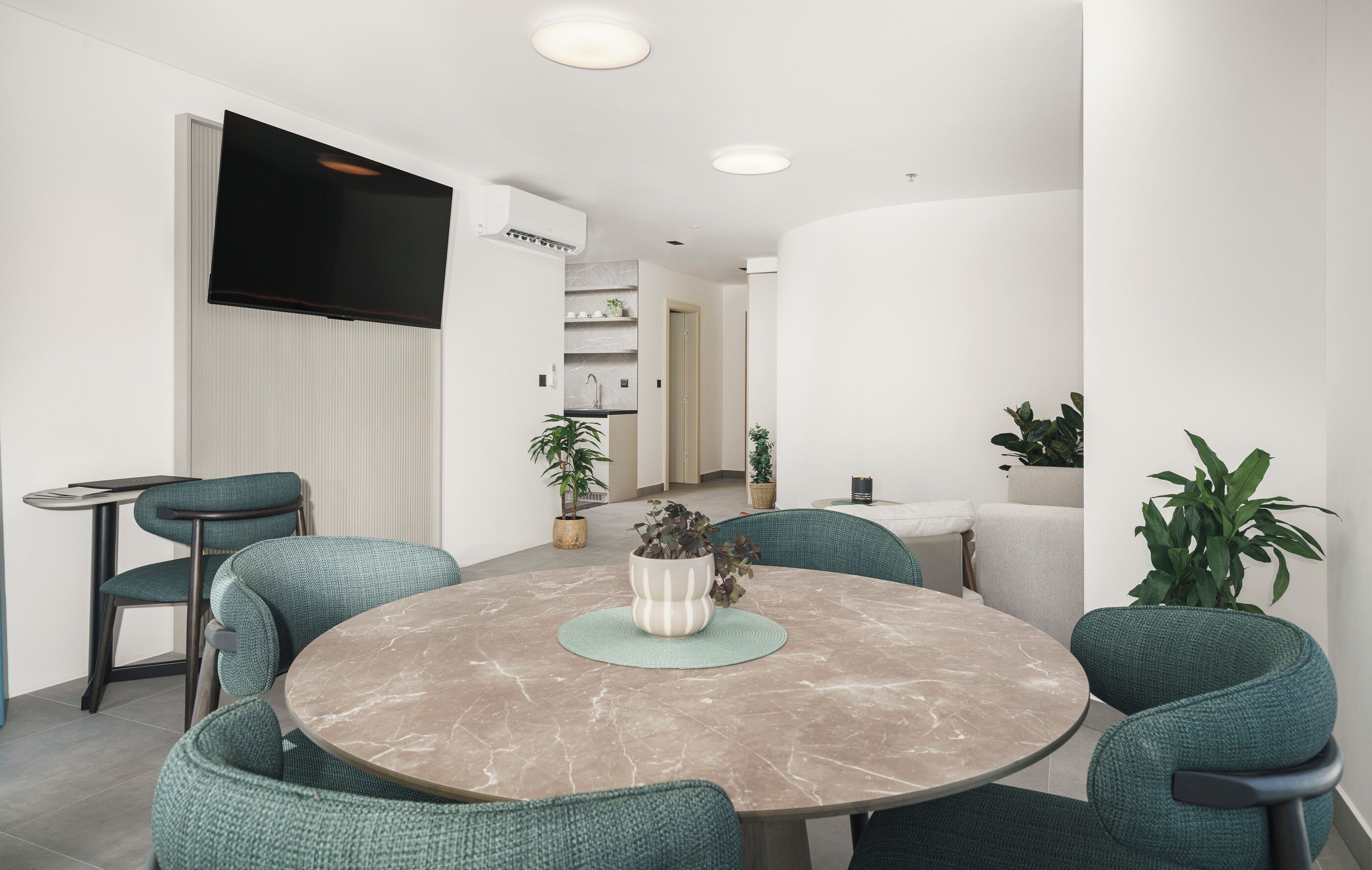 A modern dining area with a round marble table, green upholstered chairs, and potted plants.