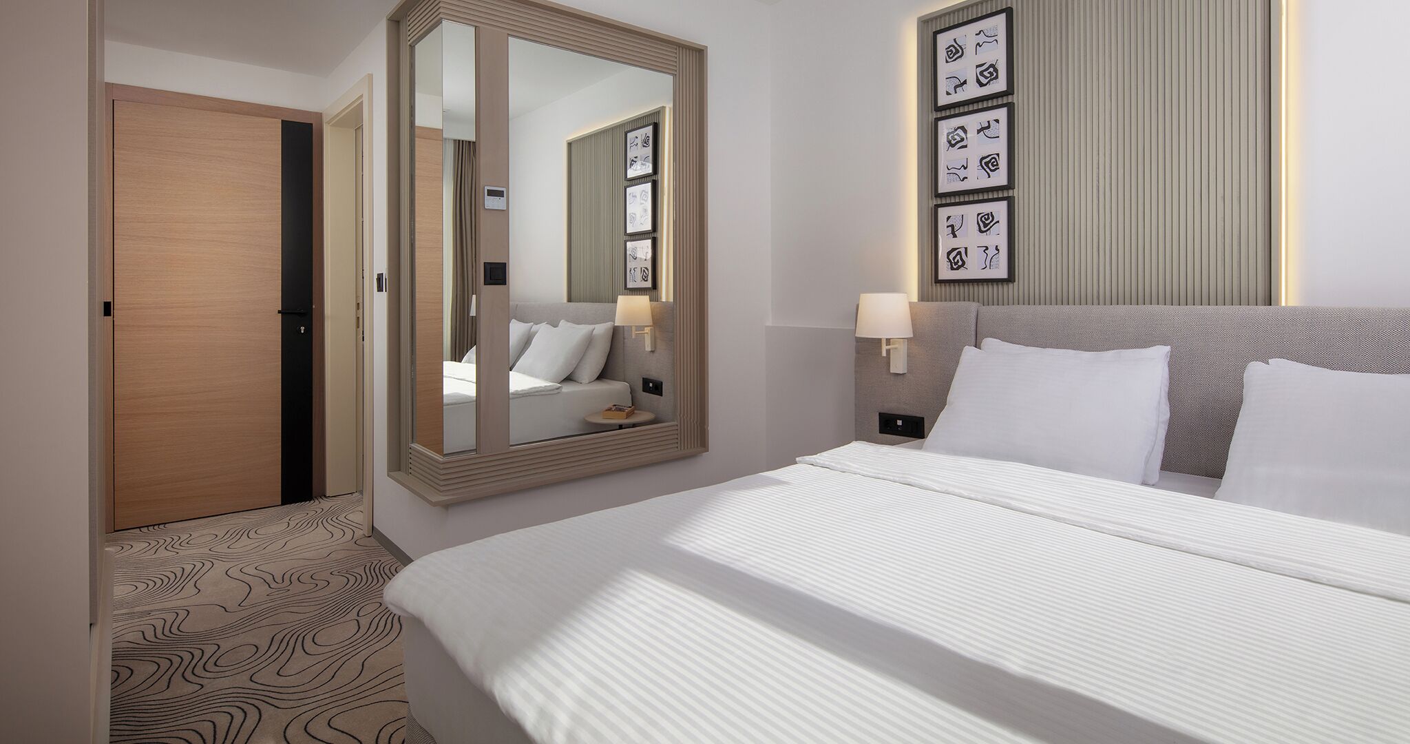 A hotel room with a large mirror, a bed with white linens, a headboard with a light fixture, and a wall with framed artwork.