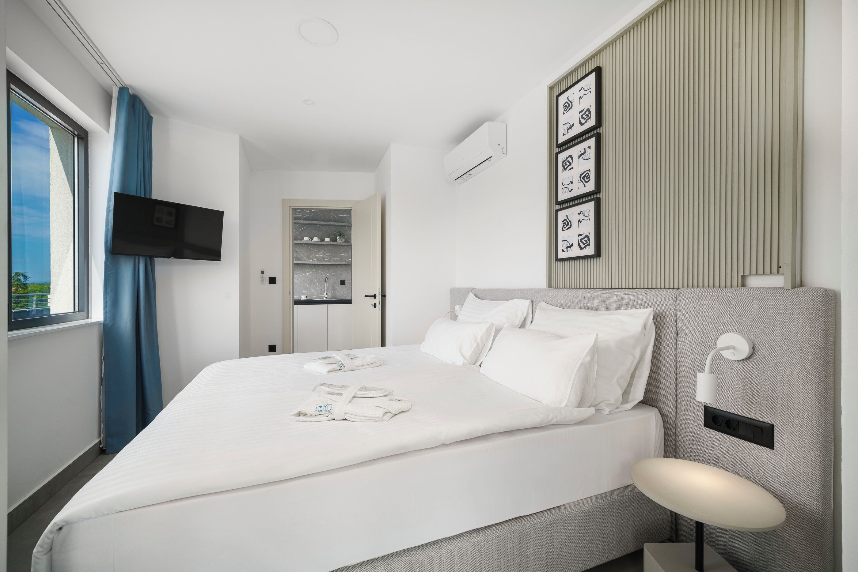 A modern hotel room with a large bed, a flat-screen TV mounted on the wall, a bedside table with a lamp, and a view of the outdoors through a window with blue curtains.