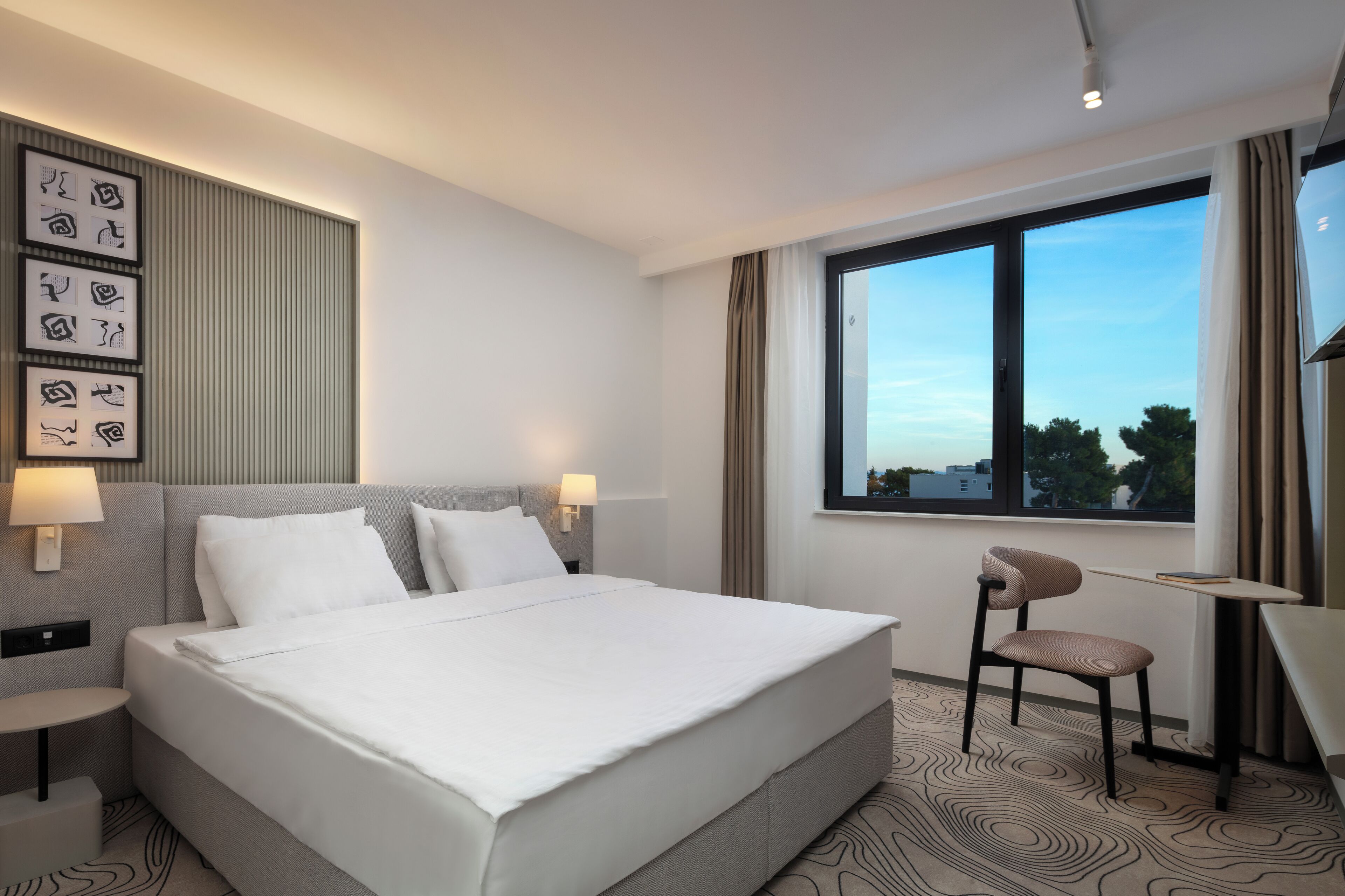 A modern hotel room with a large bed, a desk, and a window with a city view.