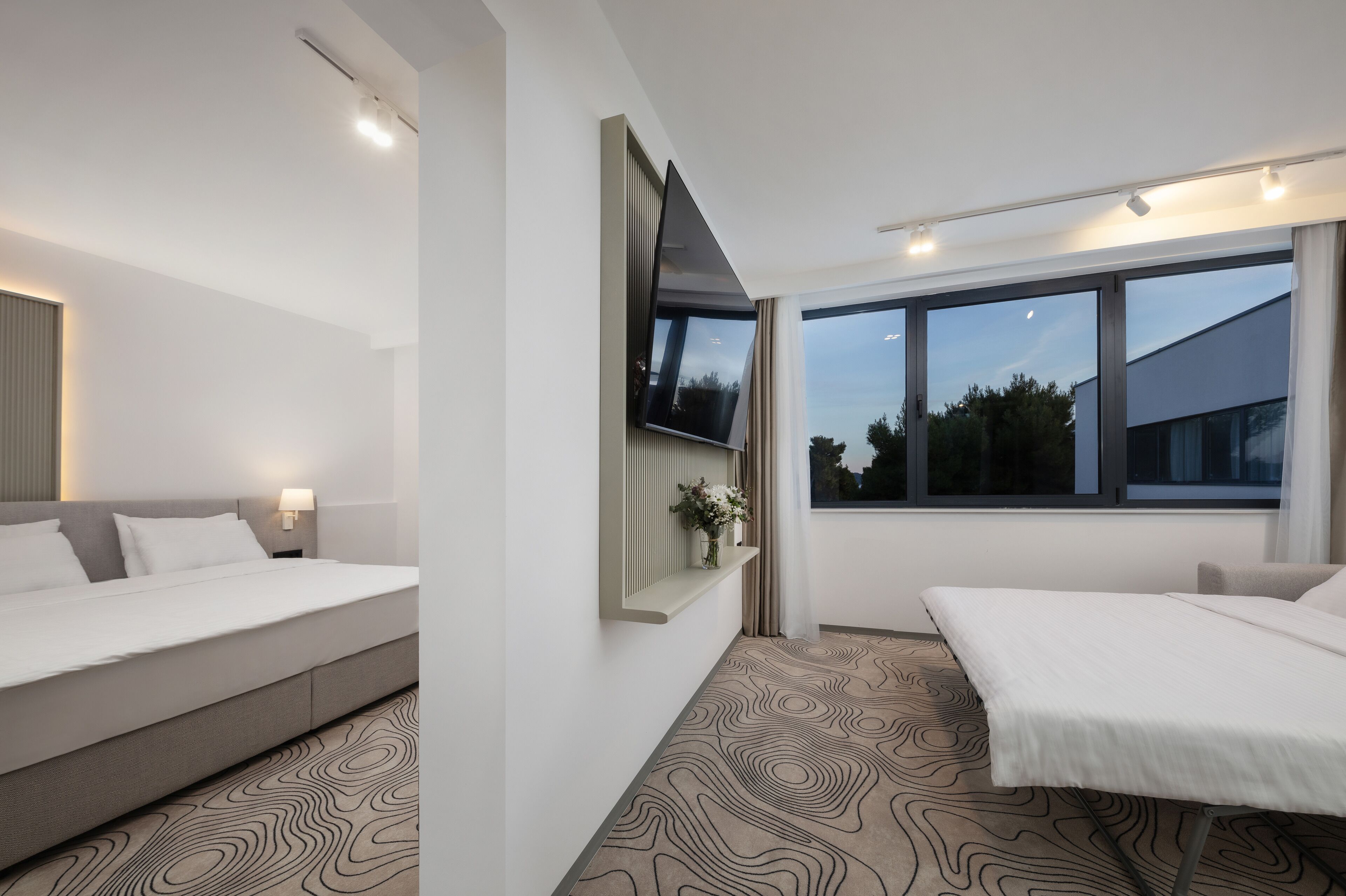 A modern hotel room with a large bed, a smaller sofa bed, a flat-screen TV mounted on the wall, and a window with a view of the outside.