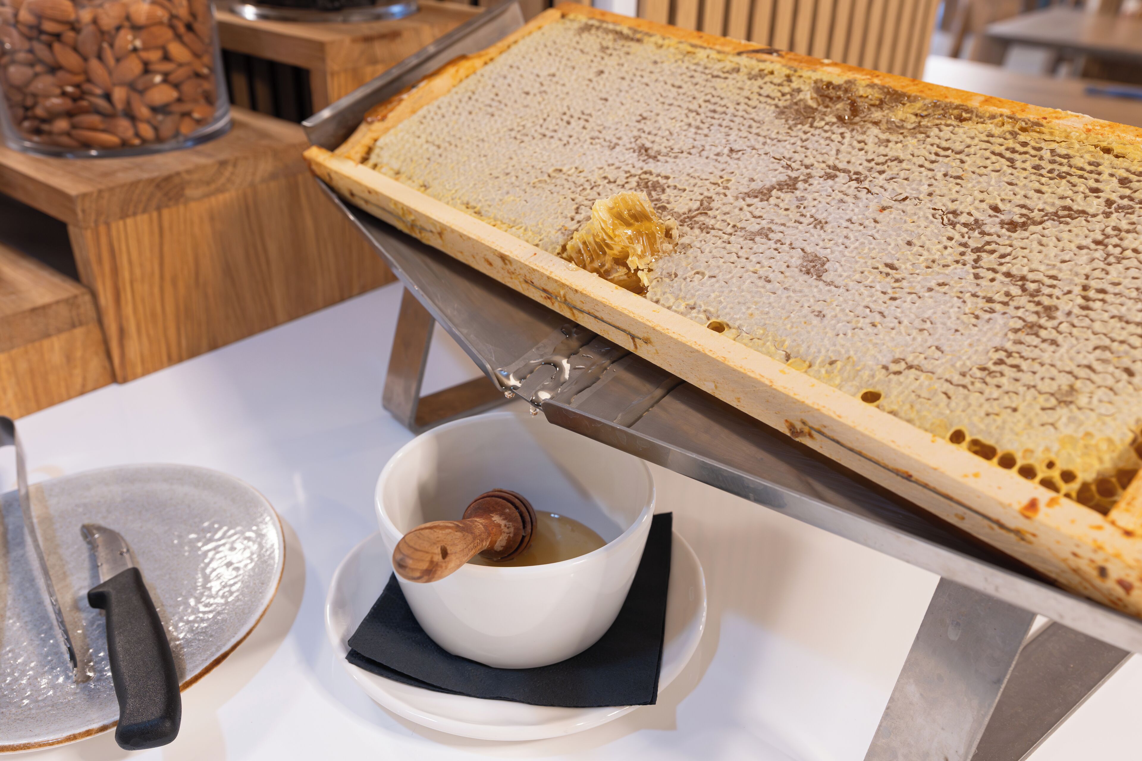 A honeycomb frame on a table with a knife and a bowl of honey.
