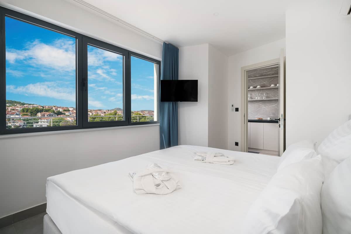 A hotel room with a large bed, a TV mounted on the wall, and a view of a cityscape through a large window.