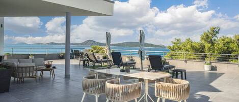 Una terraza al aire libre con vista al mar, que incluye una mesa, sillas, plantas en macetas y un área de estar cubierta.