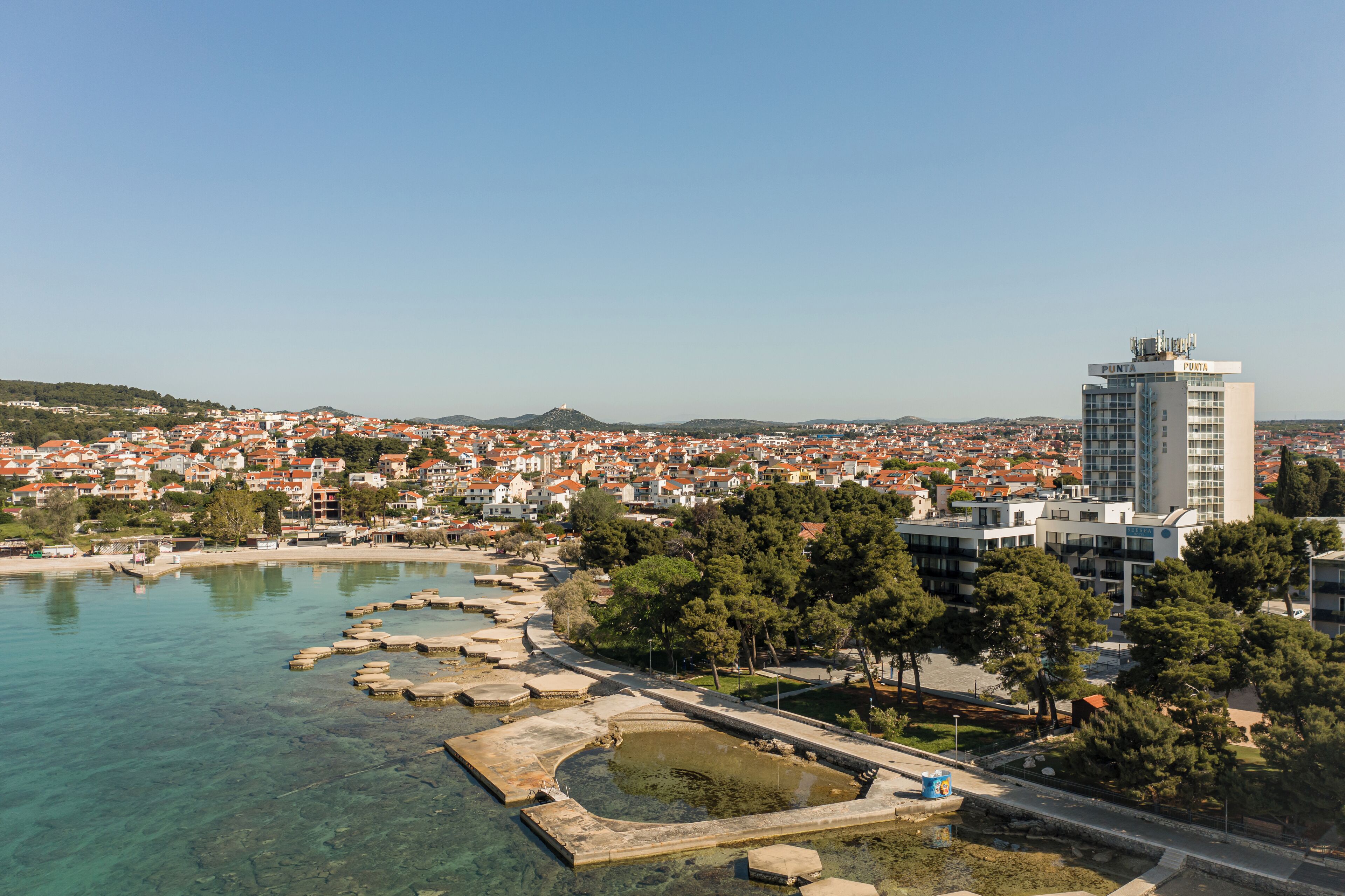 A coastal cityscape with a prominent modern building, clear waters, and a park area.