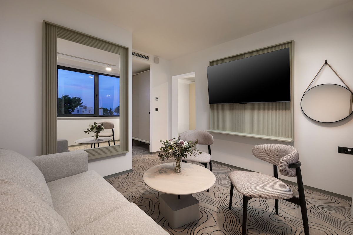 A modern living room with a sofa, two chairs, a round coffee table, and a flat-screen TV mounted on the wall.