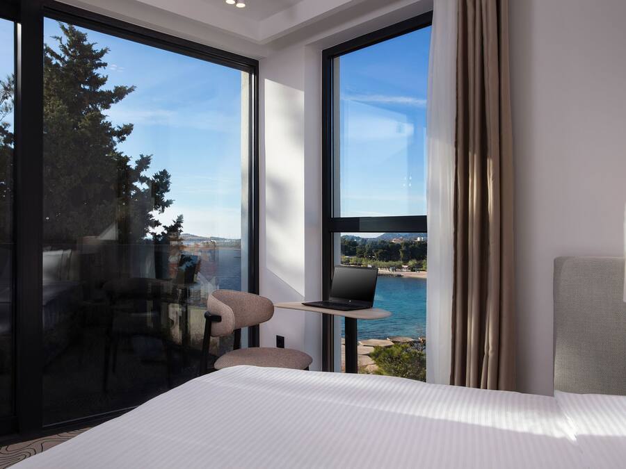 Habitación de hotel moderna con una cama grande, una laptop sobre una mesita y vistas al mar y al paisaje urbano a través de amplios ventanales.