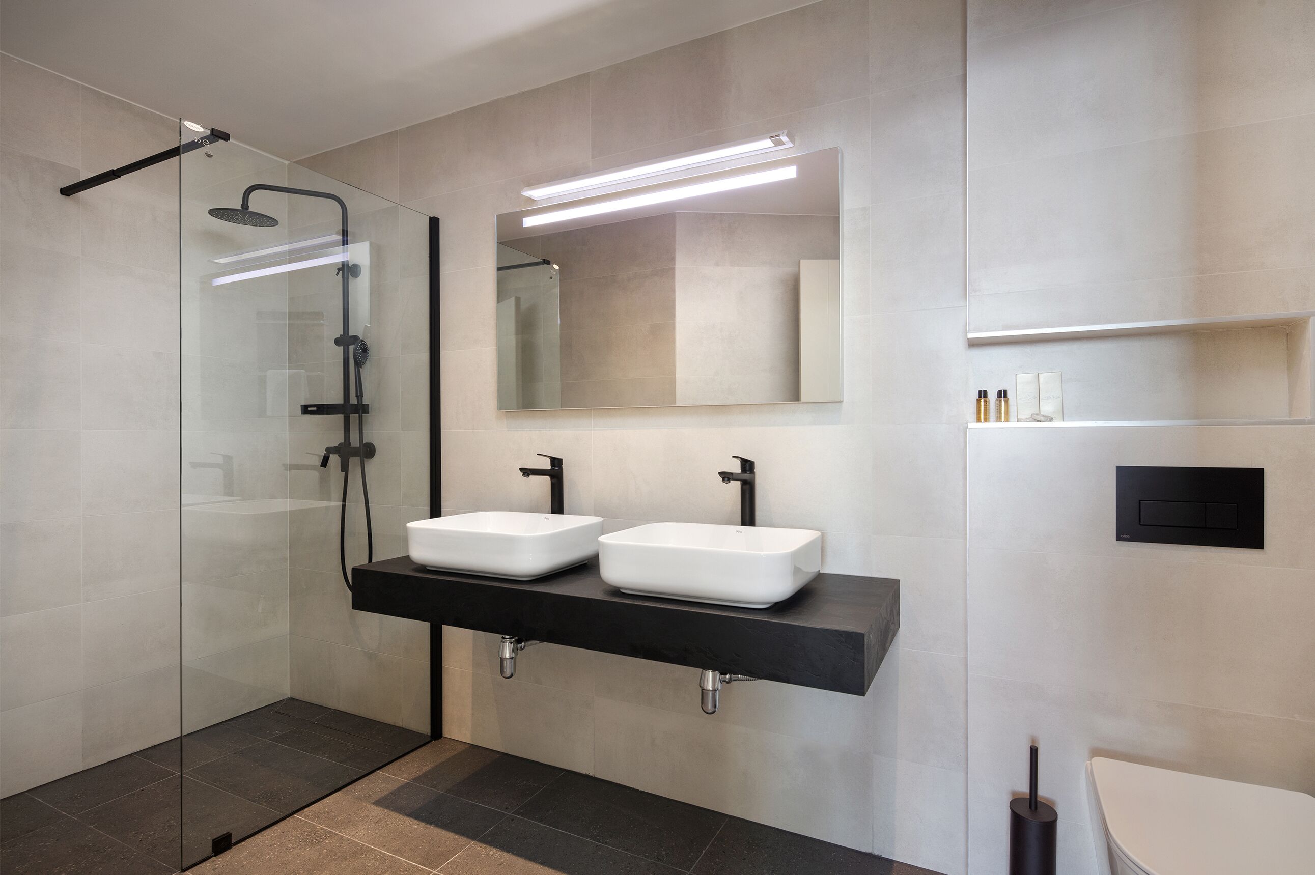 Un baño moderno con ducha envuelta en vidrio, dos lavamanos blancos con grifos negros y un espejo grande sobre los lavamanos.