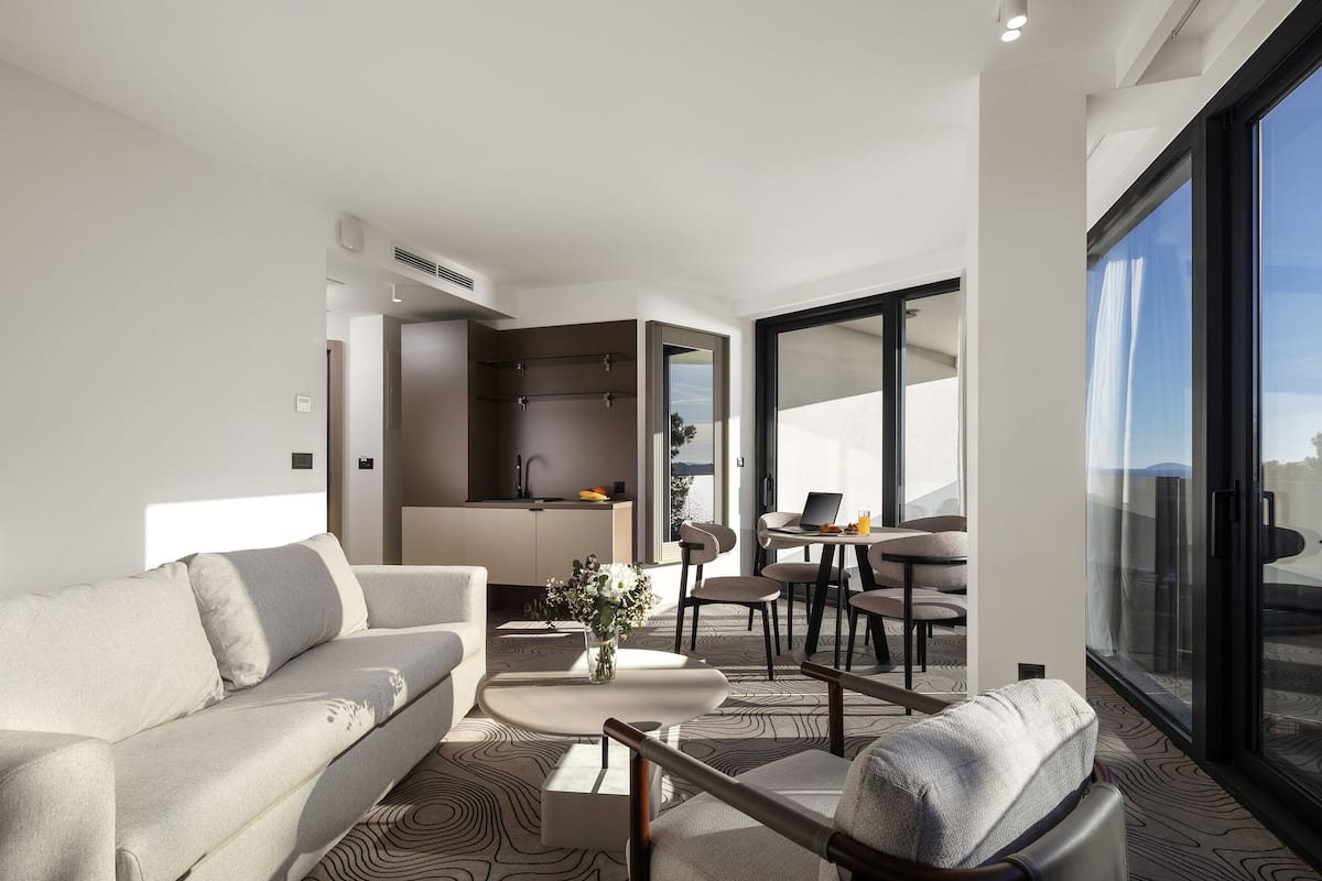 A modern hotel room with a sofa, dining area, and a large window offering a city view.