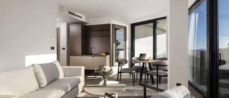 Una habitación de hotel moderna con sofá, zona de comedor y un gran ventanal con vistas a la ciudad.
