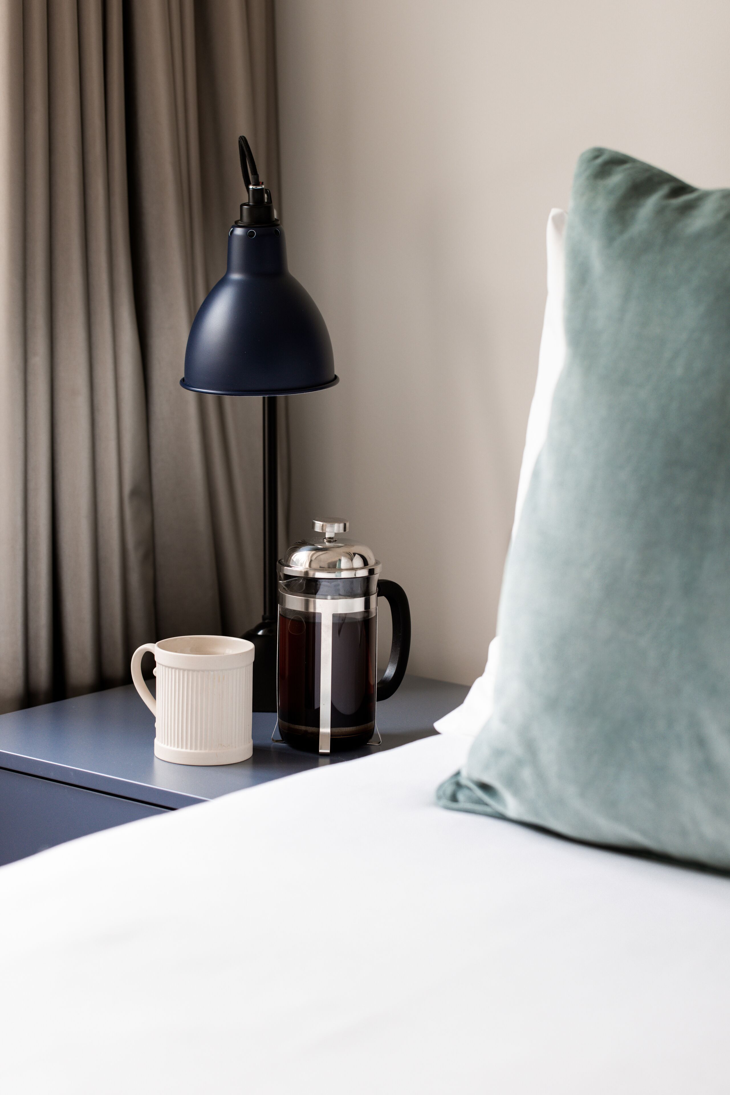 A neatly arranged hotel room with a bedside table featuring a coffee maker, a white mug, and a dark lamp.