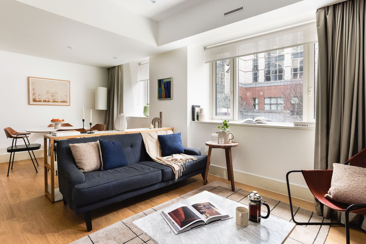 A modern living room with a dark blue sofa, a coffee table with a magazine, and a view of a building through a large window.