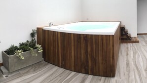 Una bañera de hidromasaje en esquina con revestimiento de madera, una jardinera de concreto con plantas, y una escalera que conduce a un nivel superior.