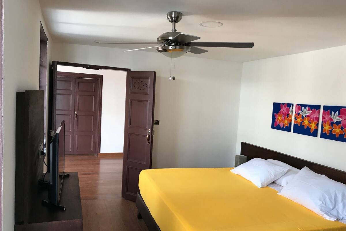 A hotel room with a bed, a ceiling fan, a television, and a door leading to another room.