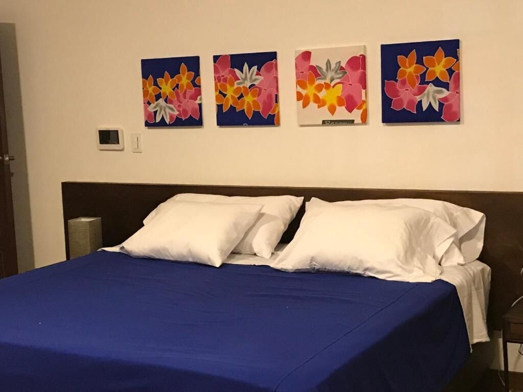 Una cama con almohadas blancas y una colcha azul, flanqueada por cuatro cuadros florales en la pared.