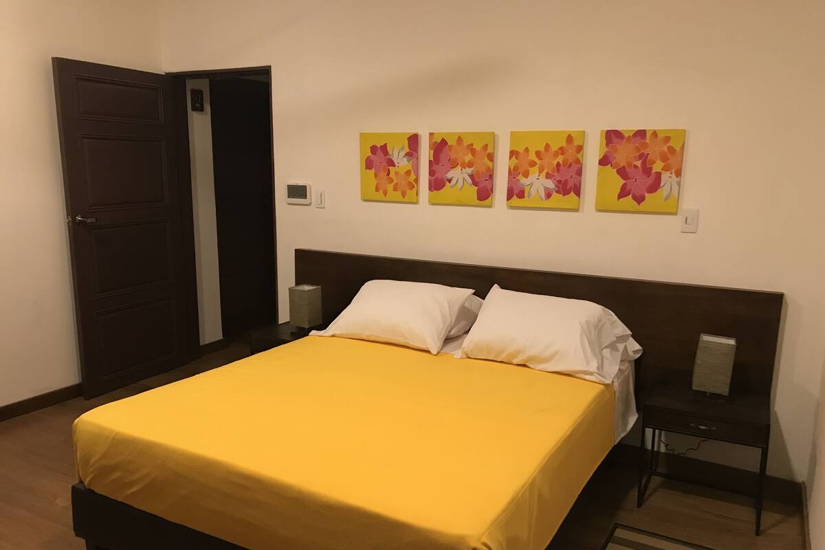 A hotel room with a bed, a nightstand, a wall clock, and floral artwork.