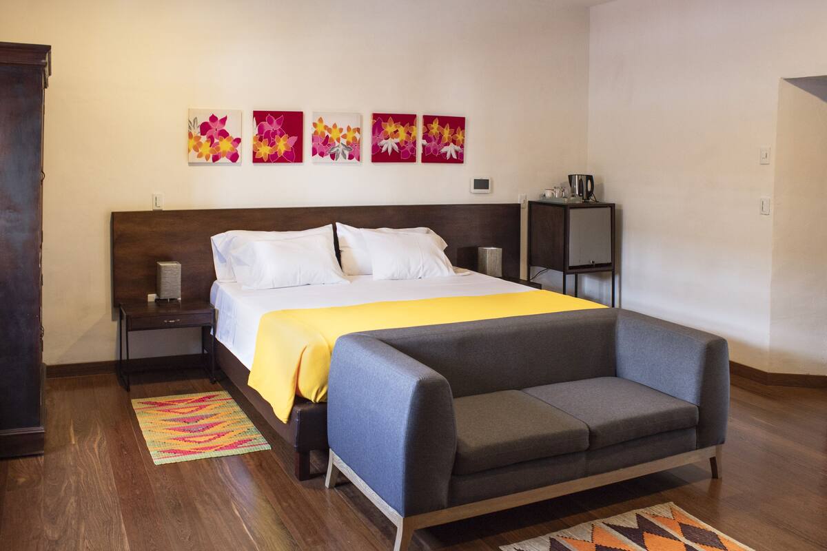 A hotel room with a bed, a sofa, a nightstand, a bedside table, a coffee maker, and floral wall art.
