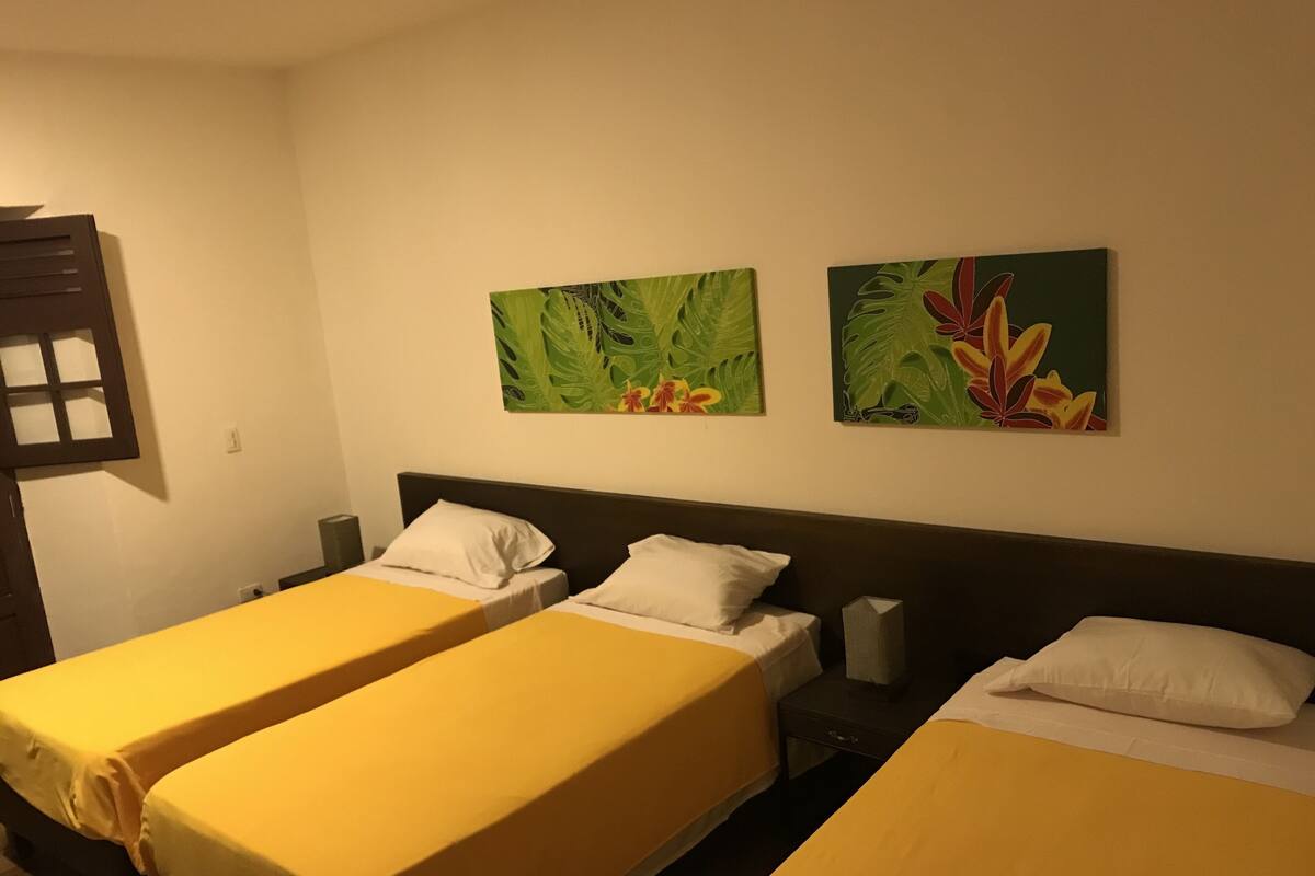A hotel room with two single beds, each with a yellow bedspread and white pillows. There are two wall-mounted paintings of tropical leaves and flowers.