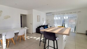 A modern dining area with a white table and chairs, a wooden bar counter, and a sofa with cushions. - L'eden des Aspres (Thuir)