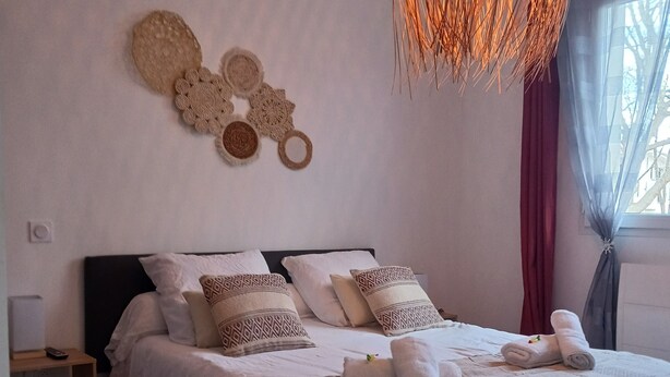 A neatly made bed with white linens and patterned pillows, a woven wall hanging, and a large, textured hanging light fixture. - L'eden des Aspres (Thuir)