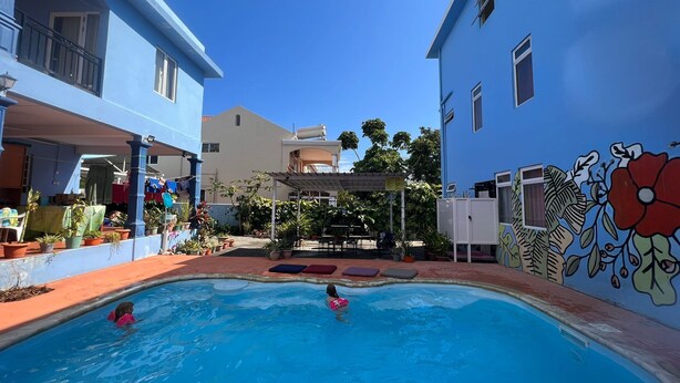 A swimming pool with two people in the water, surrounded by potted plants and a covered patio area. - Bob Apartments (Calodyne)
