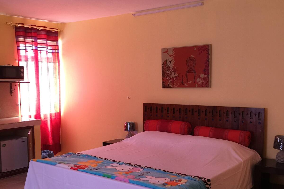 A bedroom with a bed, a ceiling fan, a desk with a microwave, and a red curtain.