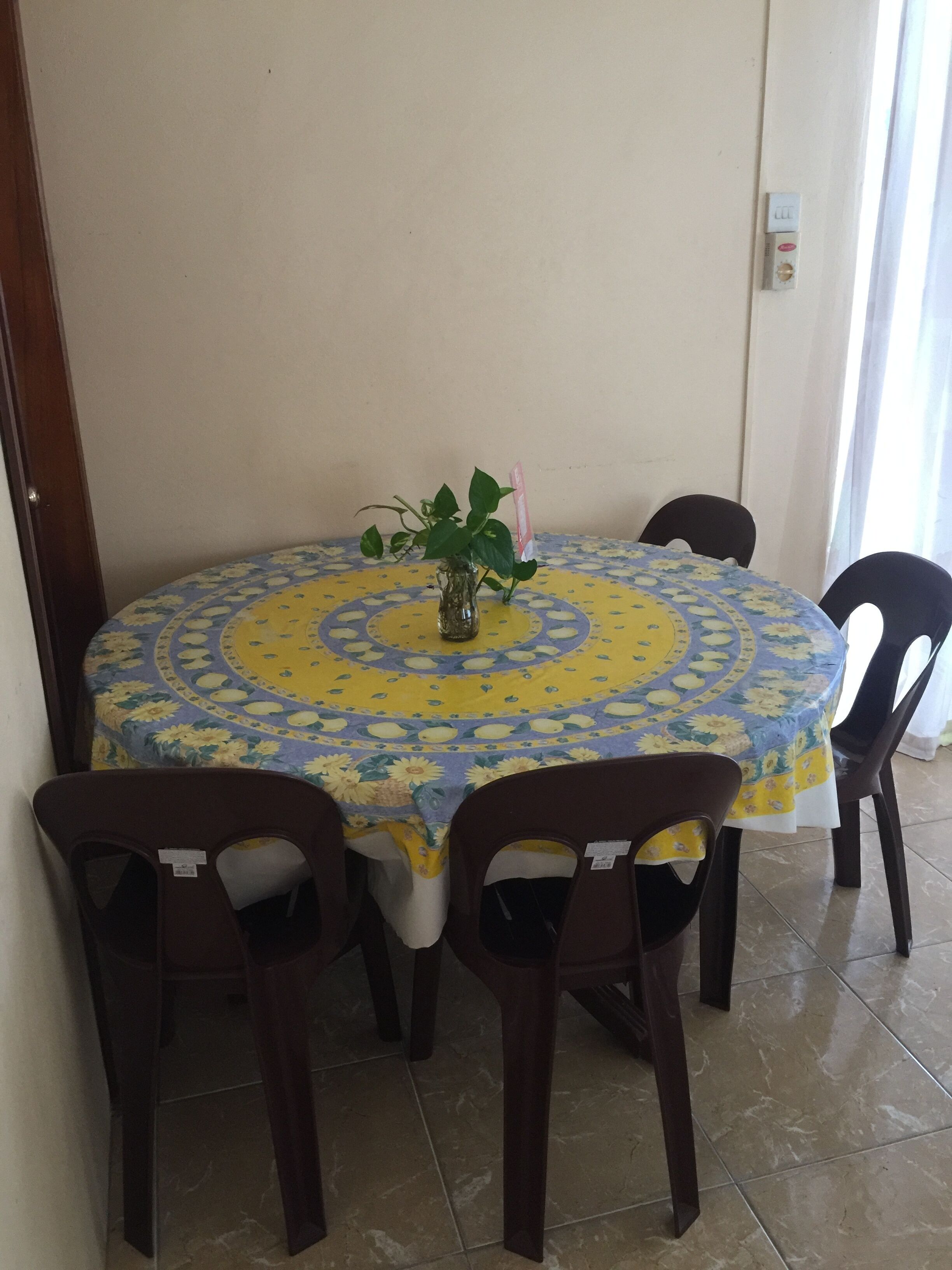 A round dining table with a floral tablecloth, surrounded by four chairs. A potted plant is placed in the center of the table.