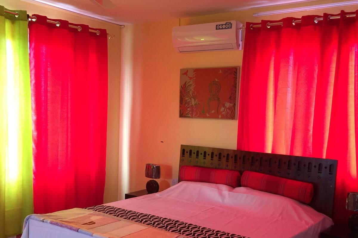 A bedroom with a bed, a ceiling fan, red curtains, an air conditioning unit, and a small table with a lamp.