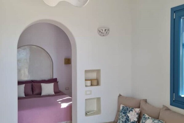 A bedroom with a bed, an arched doorway, a small shelf, and a window with a blue frame.