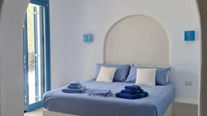 Une chambre à coucher avec un lit, une décoration aux couleurs bleu et blanc, une grande porte en arc de cercle et une vue sur l’extérieur, visible à travers cette porte.