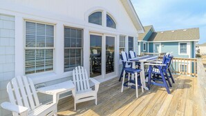 A covered deck with white and blue Adirondack chairs, a wooden table, and a glass door leading to a patio area. - Sun Realty gorgeous Nags Head home across the street from Jockey's Ridge (Nags Head)