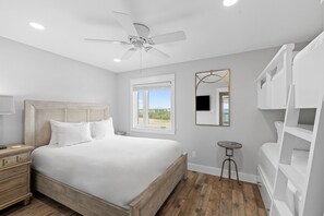 Un dormitorio con cama, ventilador de techo, ventana, mesita de noche y una estantería blanca.