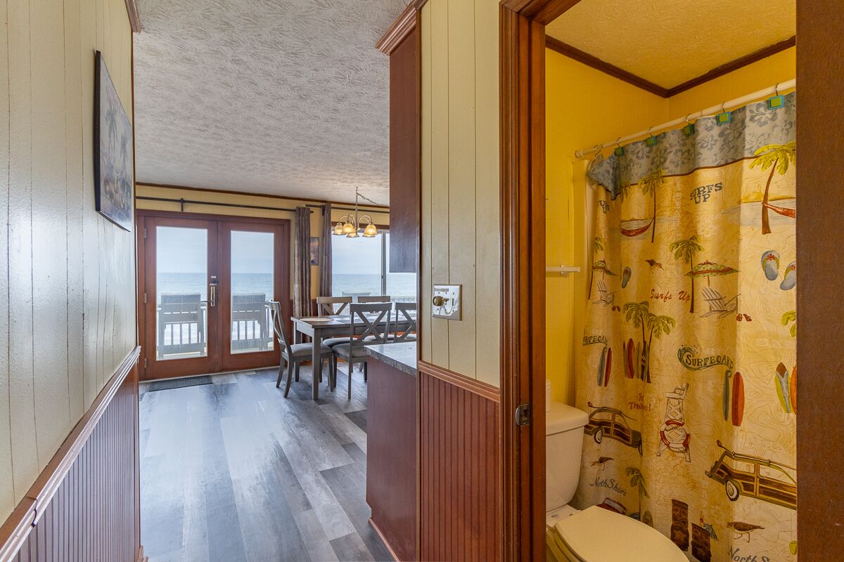 Un couloir mène à une salle à manger avec vue sur l’océan, une salle de bain avec un rideau de douche sur le thème du surf, et un plancher en bois.
