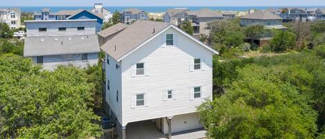 Une maison blanche à deux étages avec un garage, entourée de verdure et d’autres maisons, qui offre une vue sur une plage.