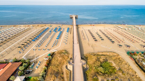 An aerial view of a beach with rows of beach chairs and umbrellas, a pier extending into the water, and a parking area with multiple levels. - Mobile Home Roma, Camping Paradiso, Viareggio, (Viareggio)