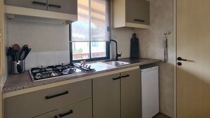 A compact kitchen with a gas stove, sink, and dishwasher. There is a window providing natural light and a view of the outside. - Mobile Home Roma, Camping Paradiso, Viareggio, (Viareggio)