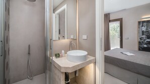 A modern bathroom with a glass-enclosed shower, a round sink, and a bedside table with a book.
