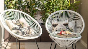 Two white wicker chairs with a glass table between them, a glass of red wine, and a plate of cheese and fruit.
